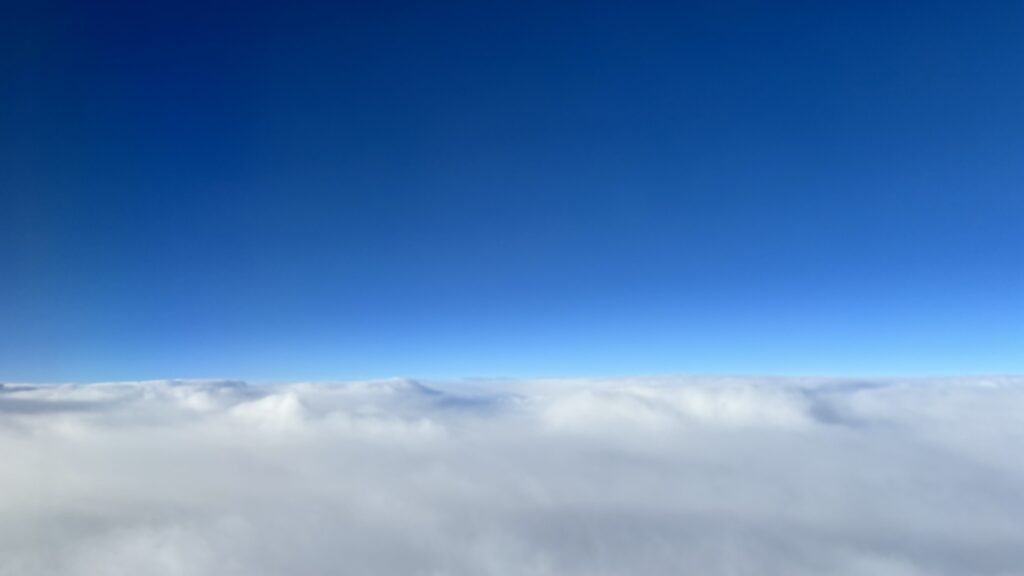 A photo taken from the window of an airplane, showing a thick, white cloud layer that looks like a mountainous landscape. Above is a clear blue sky.