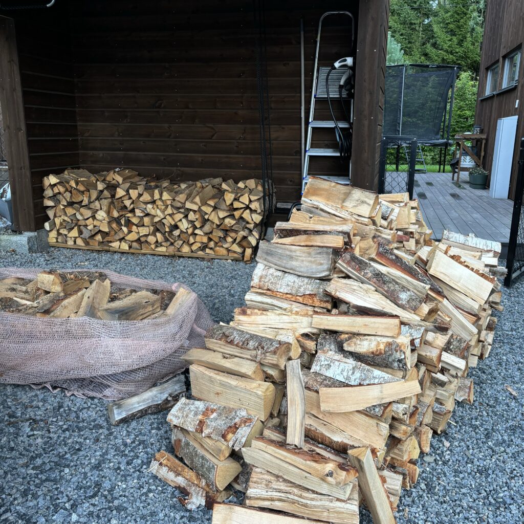 A carport with one stack and two more piles of firewood. 