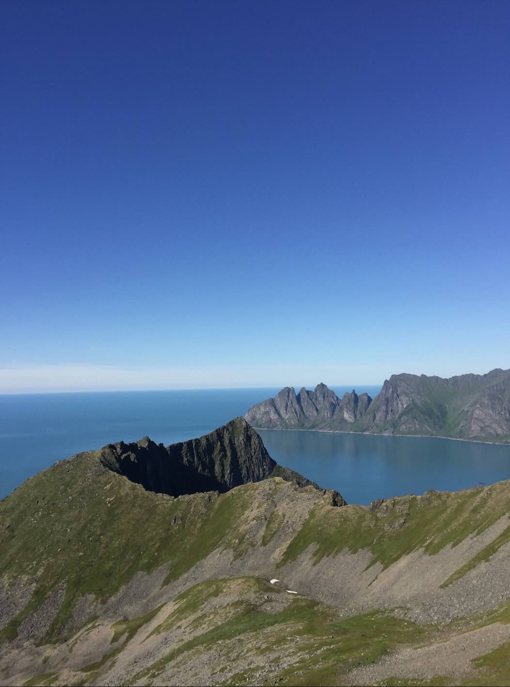 Photo of a mountainous landscape, with green terrain, rocky ridges, and a fjord-like bay. The sky is clear blue and there's a range of jagged peaks in the distance.