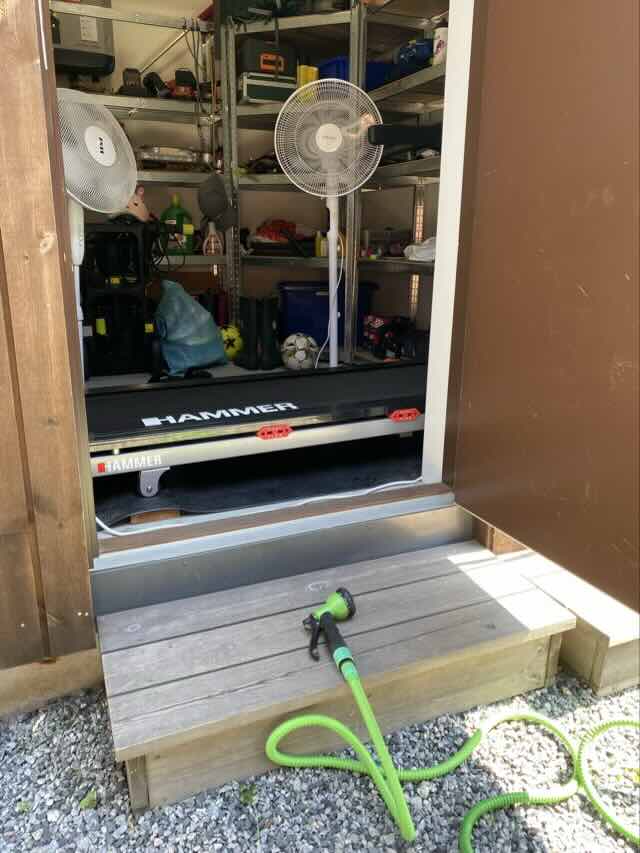 Photo of a treadmill in a shed, with two fans, an open door and a garden hose for cooling.