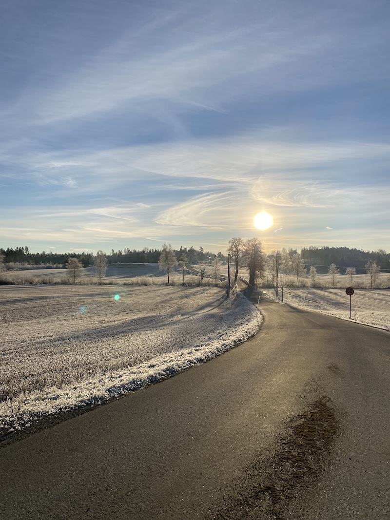 Photograph of the sun shining over a rural landscape showing a back country road going through fields covered by hoarfrost